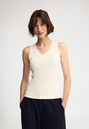 Femme aux cheveux courts et bruns portant un haut crème côtelé sans manches et un pantalon ample foncé, debout les mains dans les poches sur fond blanc.