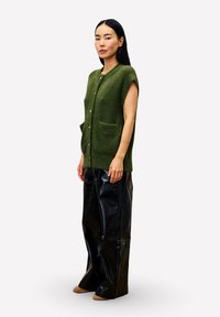 Vrouw staat in een mouwloze groene button-up cardigan, wijde zwarte glanzende broek en bruine puntige schoenen.