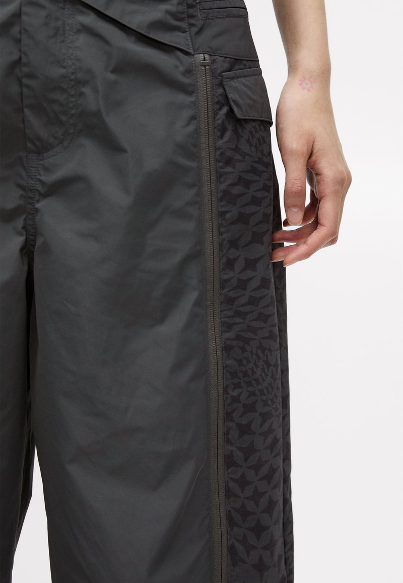 Lätta, mörkgrå byxor med en slät textur, som har ett mönstrat sidofält, dragkedjestängning och en höftficka för funktionalitet.