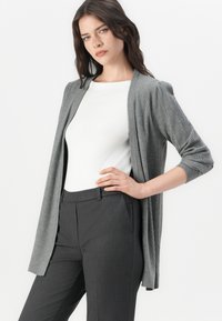 Gilet tricot gris avec texture côtelée, ouverture sur le devant et manches trois-quarts, porté sur un haut blanc et un pantalon noir ajusté.