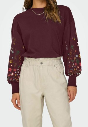 Frau trägt ein kastanienbraunes langärmeliges Oberteil mit Blumenmuster an den Ärmeln und eine hoch taillierte beige Hose mit Knopfverschluss.