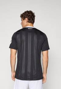 Man in black striped sports jersey with "RMCF" text on the upper back, facing away, wearing white shorts against a plain background.