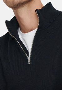 Ciemnoniebieski sweter na zamek z prążkowaną teksturą i wysokim kołnierzem, z metalowym zamkiem błyskawicznym, noszony na białej koszulce z okrągłym dekoltem.