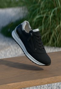 Black sneaker with a textured upper, silver accents, a side zipper, and a white sole. Designed with a sleek shape and modern materials.