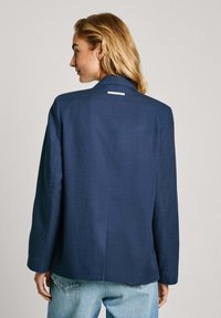 Mornarško modra blazer jakna iz lahkega materiala, z izrezanim ovratnikom, dolgimi rokavi in majhno etiketo na hrbtni strani ovratnika.