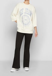 Sudadera oversize crema con logo gráfico azul, combinada con pantalones negros acampanados y zapatillas blancas. Tela suave, ajuste relajado.