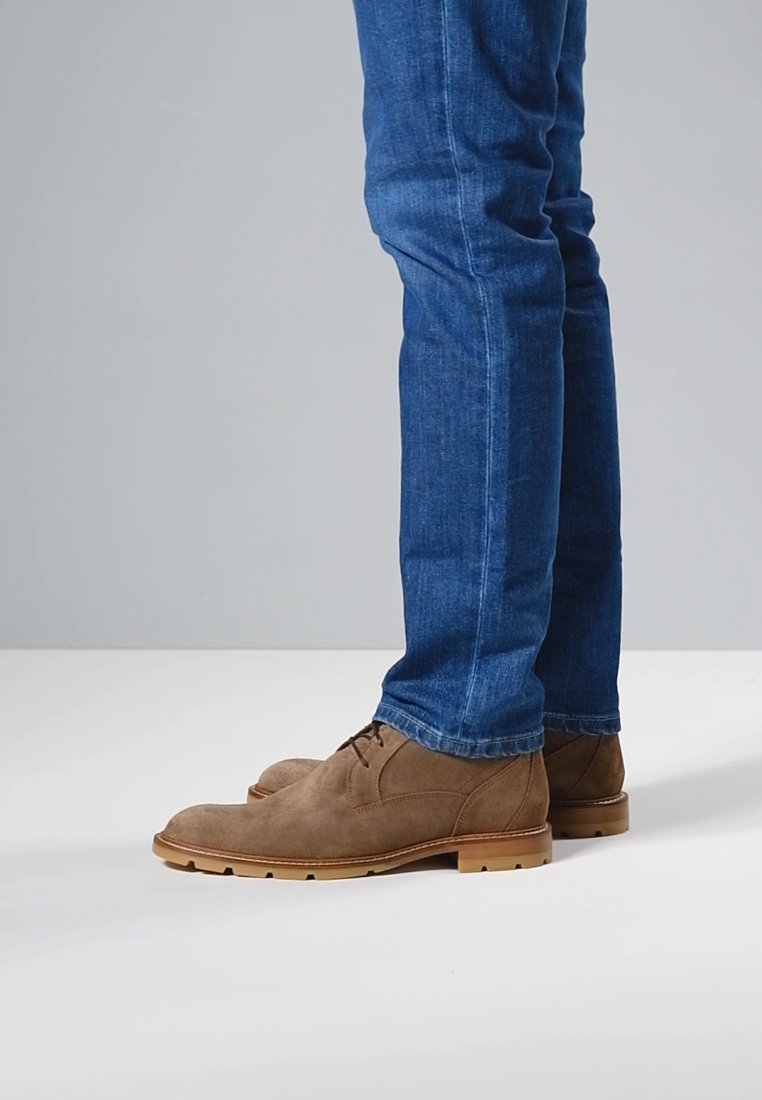 Brown suede ankle boots with a rounded toe and rubber sole, paired with fitted blue denim jeans, showcasing a casual, contemporary style.