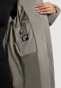 Interior de una chaqueta acolchada gris claro con un pequeño bolsillo de etiqueta negra. La tela parece suave con una textura ligeramente brillante.