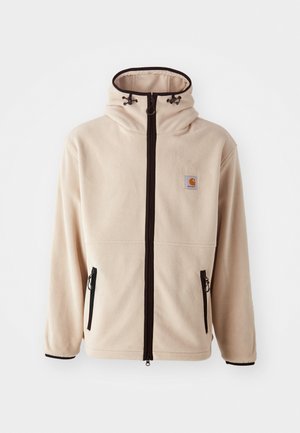Chaqueta de forro polar beige con cuello alto, cremalleras negras y bolsillos laterales. Presenta un parche con logo en el pecho y un borde negro contrastante.