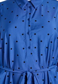 Blaue Hemdkleid mit schwarzen Tupfen, Knopfleiste vorne, Kragen und passendem Stoffgürtel, der um die Taille gebunden ist.