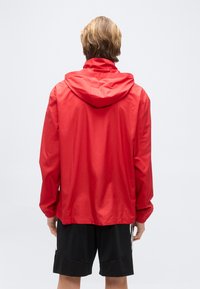 Άτομο με κοντά ξανθά μαλλιά που φοράει φωτεινό κόκκινο μπουφάν με κουκούλα και μαύρο σορτς, στέκεται με την πλάτη προς τα εμπρός σε απλό φόντο.