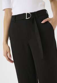 Zwarte op maat gemaakte broek met een brede pijp, voorzien van een D-vormige zilveren gesp riem en een strikdetail bij de taille. Soepele stoftextuur.