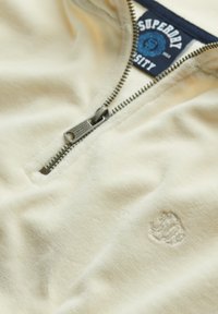 Sudadera crema de media cremallera hecha de tejido suave, con cremallera metálica, logotipo en relieve y una etiqueta azul con el nombre de la marca en el cuello.