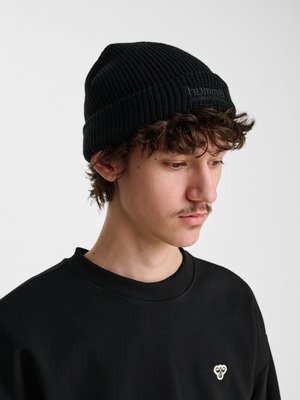 Junger Mann mit lockigem Haar, der eine schwarze Strickmütze und ein schwarzes Sweatshirt mit kleinem weißem Logo trägt, schaut nach unten und zur Seite.