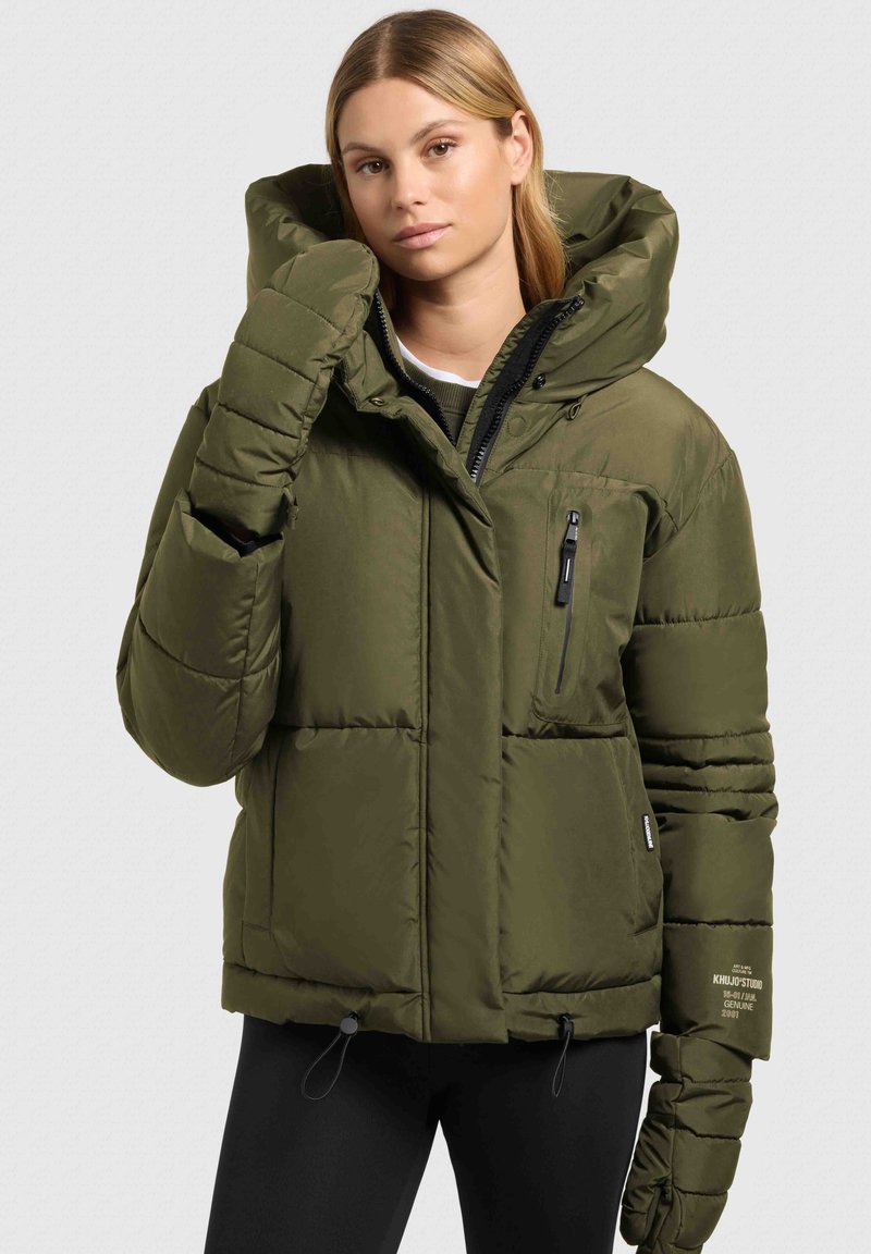 Doudoune vert olive avec une grande capuche, une poche poitrine zippée, des poignets élastiques et un ourlet à cordon de serrage ; texture matelassée sur l'ensemble.