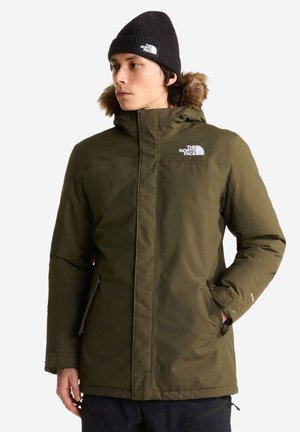 Mann trägt olivgrüne, isolierte Parka mit fellbesetzter Kapuze und schwarzer Strickmütze, Hände in den Taschen, schaut nach links vor einfarbigem Hintergrund.