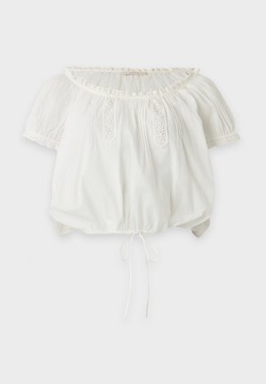 Femme debout portant un chemisier blanc épaules dénudées, un pantalon blanc taille haute avec cordon et des sandales marron à talons sur fond uni.