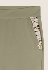 Pantalones de chándal en verde oliva claro con una textura suave, que cuentan con un bolsillo estampado en beige, negro y blanco. Etiqueta de marca en tono dorado en el lateral.