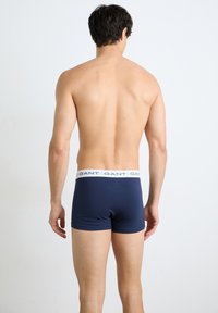 Άνδρας που στέκεται με την πλάτη, φορά σκούρα μπλε μποξεράκια με λευκή ζώνη με το λογότυπο "GANT" σε απλό λευκό φόντο.