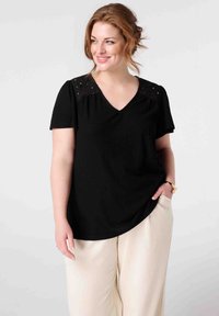 Haut noir à col en V avec manches courtes, présentant des détails en dentelle sur les épaules, en tissu texturé, associé à un pantalon large de couleur crème.