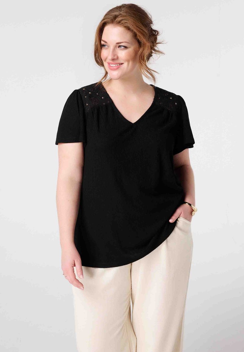 Haut noir à col en V avec manches courtes, présentant des détails en dentelle sur les épaules, en tissu texturé, associé à un pantalon large de couleur crème.