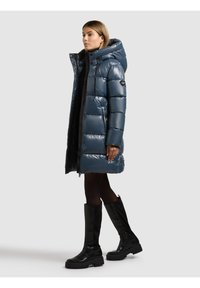 Blå vadderad jacka med glänsande finish, utrustad med huva, dragkedja framtill och förlängd, quiltad design. Bärs med knähöga svarta stövlar.