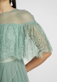 Femme portant une robe en dentelle vert menthe avec un empiècement transparent à pois et des manches froufrou, assortie à une grande boucle d’oreille en forme de goutte.