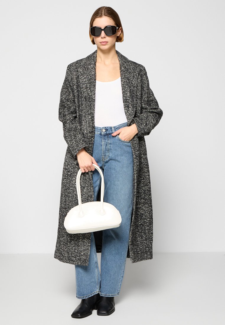 Vrouw met zwarte zonnebril, lange grijze gemusterde jas, witte top, blauwe spijkerbroek, zwarte schoenen, en een witte handtas.