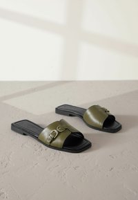 Sandales à enfiler vert olive avec une finition brillante, bout ouvert carré et un détail décoratif de boucle argentée sur la bride. Semelle noire.