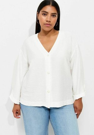 Vrouw met stijl donker haar die creooloorbellen draagt, een witte blouse met knopen en V-hals, en lichtblauwe spijkerbroek, staand voor een witte achtergrond.