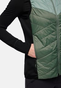Gilet imbottito verde con una maglia a maniche lunghe nera. Presenta tasche laterali con zip e un tessuto morbido e texturizzato.