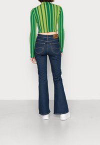 Top corto a righe verde e gialle abbinato a jeans svasati blu scuro. Include un'etichetta con il logo Lee e scarpe bianche.