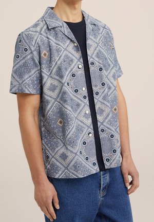 Uomo che indossa una camicia blu a maniche corte con motivi sopra una t-shirt nera e jeans blu, in piedi davanti a uno sfondo semplice.
