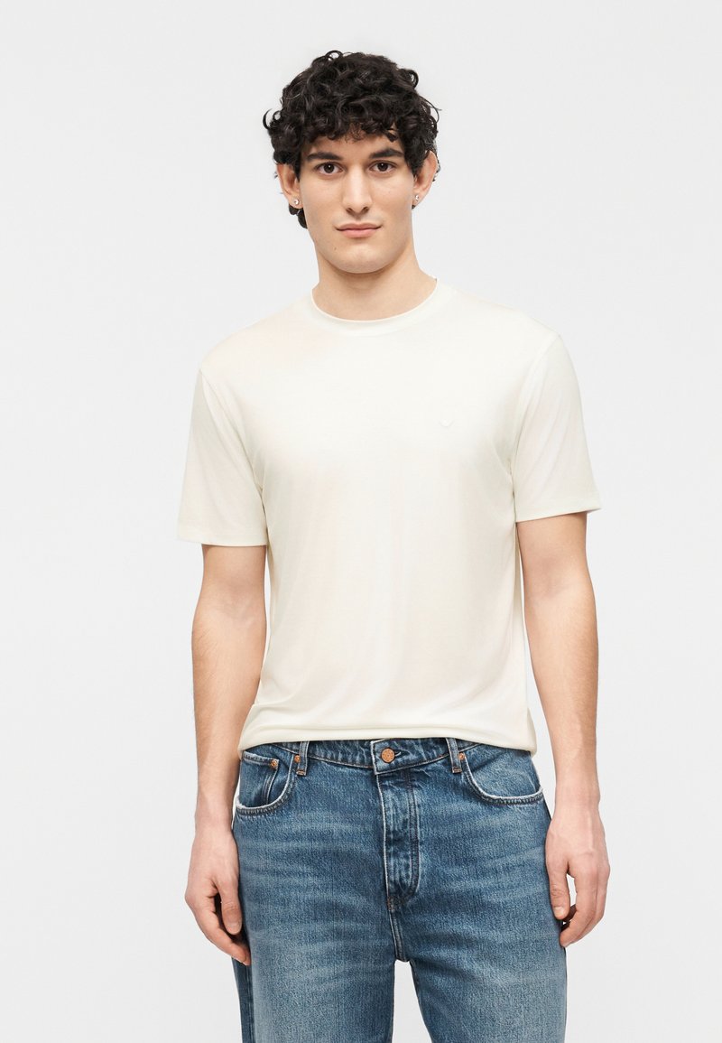 Νεαρός άνδρας με κατσαρά σκούρα μαλλιά, φοράει α πλό κοντομάνικο μπλουζάκι εκρού και μπλε τζιν, στέκεται μπροστά σε ουδέτερο φόντο.