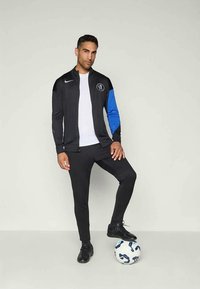 Schwarze Sportjacke mit blauem Ärmel, über einem weißen Shirt, kombiniert mit schwarzen Hosen und schwarzen Fußballschuhen. Ein Fußball liegt zu seinen Füßen.