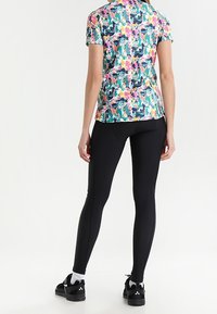 Mujer de pie de espaldas, vistiendo una camisa de manga corta con estampado abstracto y colorido, leggings negros, calcetines blancos y zapatillas negras.