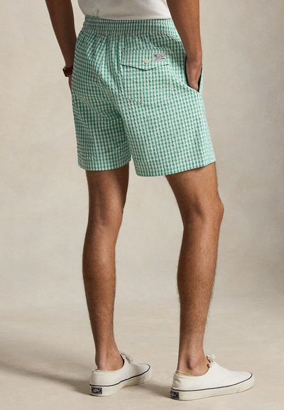 Mees, kes kannab rohevalgete ruuduliste pükste tagataskuga, valged madalad tossud, käed taskutes, seisab neutraalsel taustal.