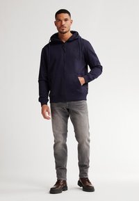 Sudadera con cremallera de color Navy con capucha ajustable, bolsillos laterales y un parche con el logo, combinada con jeans grises y botas de cordones marrones.