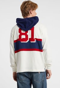 Witte hoodie met een marineblauwe capuchon en een groot rood nummer "81" op de achterkant. Heeft horizontale strepen in marineblauw en rood over de schouders.