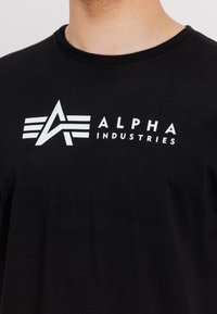 Sort bomulds T-shirt med et hvidt logo på brystet med teksten "ALPHA INDUSTRIES" og vinklede designelementer.