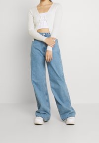 Bílý žebrovaný kardigan přes bílý crop top, doplněný světle modrými širokými džíny a bíl�ými teniskami. Jednoduchý design a ležérní střih.