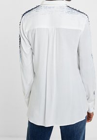 Vit skjorta med lång ärm och vik i ryggen, med textaccenter i blått längs axlarna och dekorativt trim vid halsringningen.