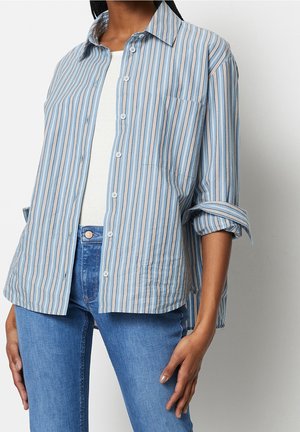 Person wearing a light blue, white, and beige vertical striped button-up shirt over a white top with blue denim jeans.