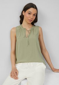 Haut sans manches de couleur vert clair avec un col à volants et un détail à nouer. Tissu lisse, coupe décontractée, associé à un pantalon blanc.