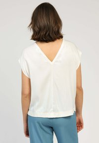 Femme avec les cheveux bruns courts portant un haut sans manches blanc à dos en V et un pantalon turquoise, se tenant devant un fond uni.