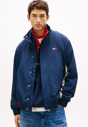 Ung mand iført lagdelte jakker i marineblå, rød og hvid med Tommy Hilfiger-logo, kombineret med blå jeans, mod en ensfarvet hvid baggrund.