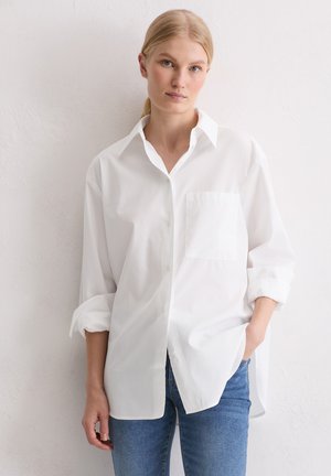 Biała koszula zapinana na guziki z klasycznym kołnierzykiem, długimi rękawami, pojedynczą kieszenią na piersi i luźnym krojem, zestawiona z jasnoniebieskimi dżinsami.