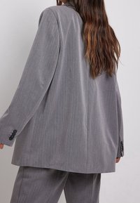 Blazer gris sur mesure avec une coupe décontractée, fermeture à un bouton et une texture subtile rayée. Présente des boutons noirs sur les poignets.