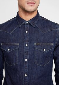 Granatowa koszula z guzikami z wywiniętym kołnierzykiem, dwiema kieszeniami na piersi, kontrastowymi szwami i srebrnymi zatrzaskami.