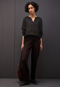 Bluse mit schwarz-gesticktem Leopardenmuster, V-Ausschnitt, langen Ärmeln; kombiniert mit braunen, weiten Hosen mit Fronttaschen. Schwarze Schuhe und eine braune Tasche.